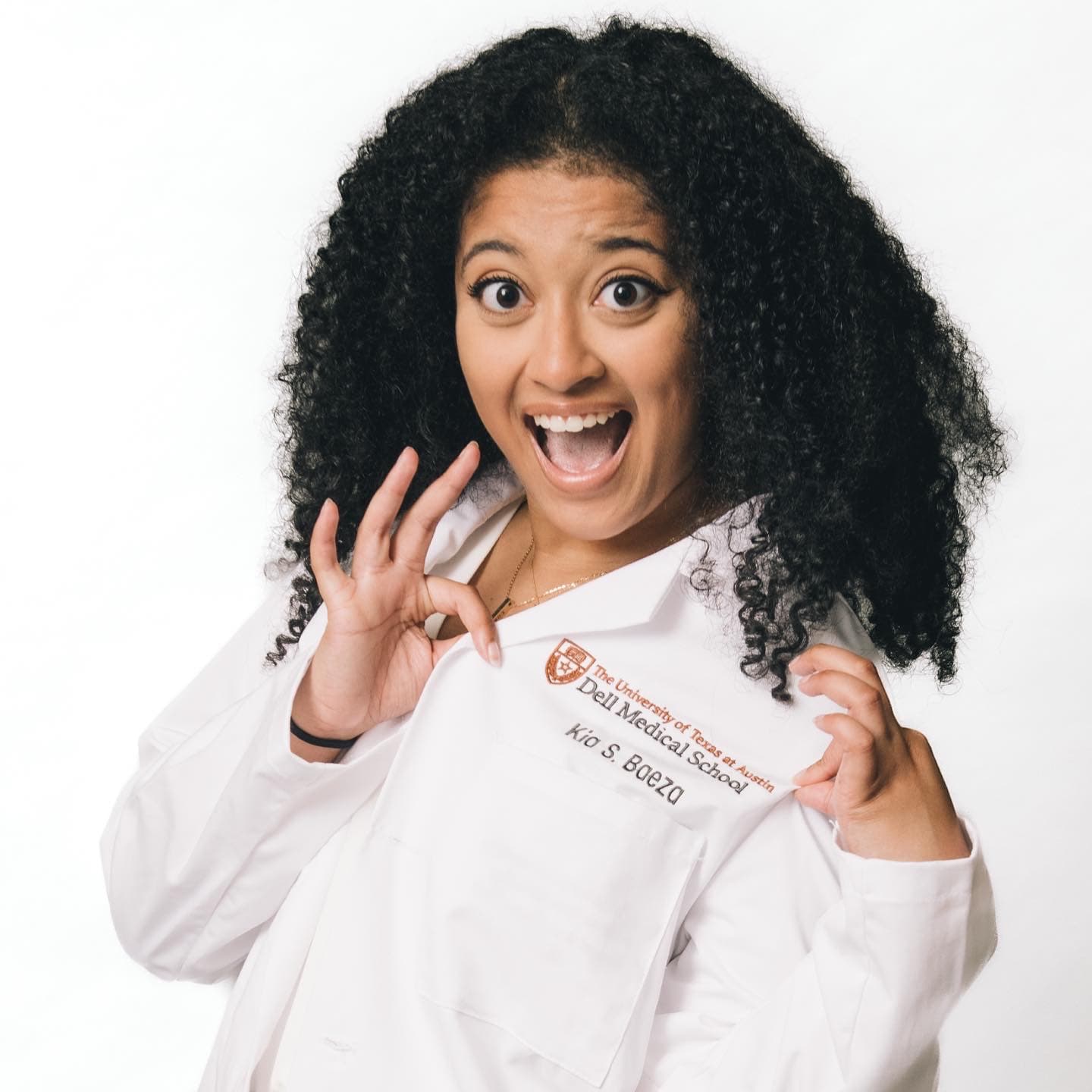 Excited woman with curly hair proudly displays her Dell Medical School white lab coat.