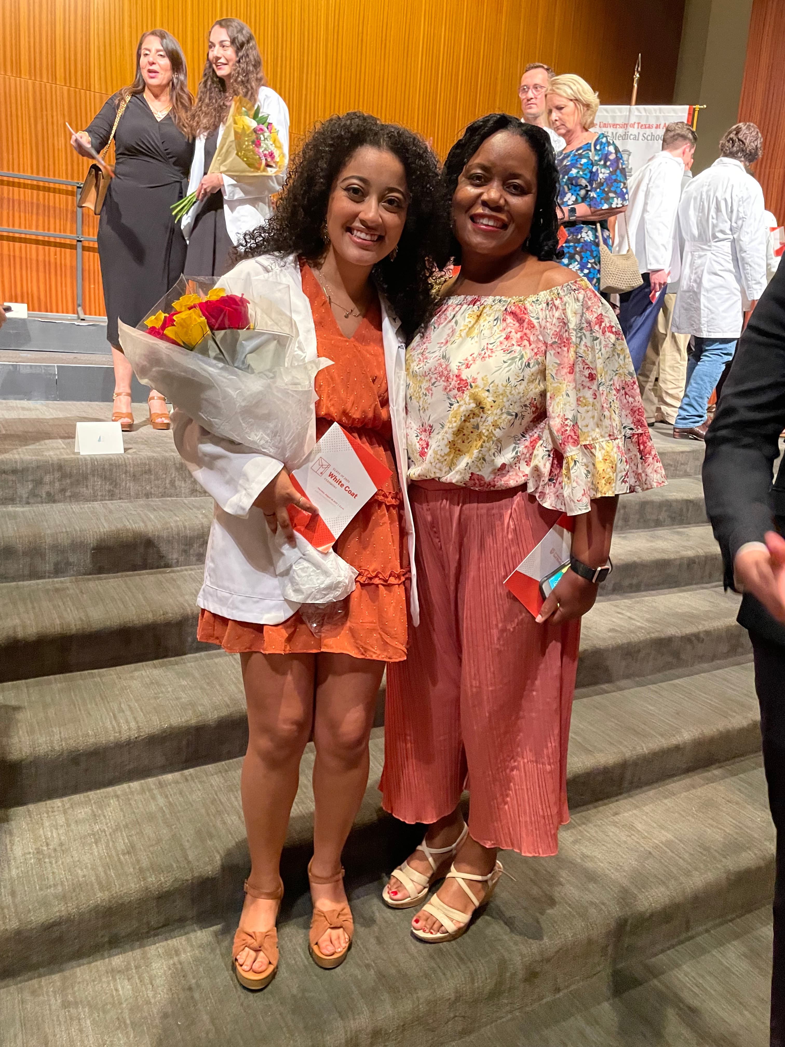 Two women smile on stairs at a White Coat Ceremony, one holding flowers and program.