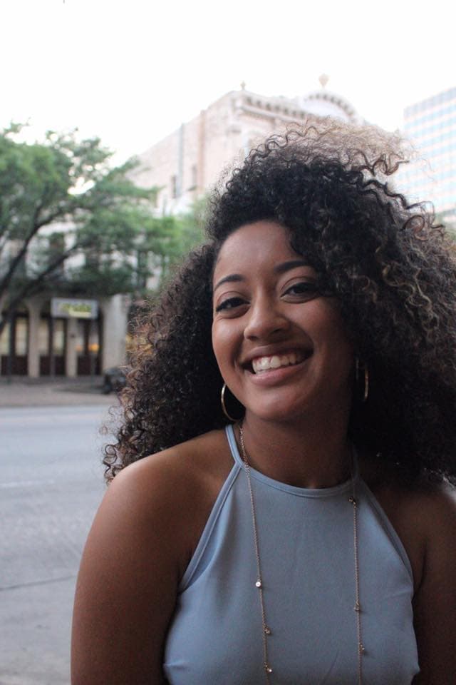 Smiling woman with voluminous curly hair and a blue halter top against a city background.