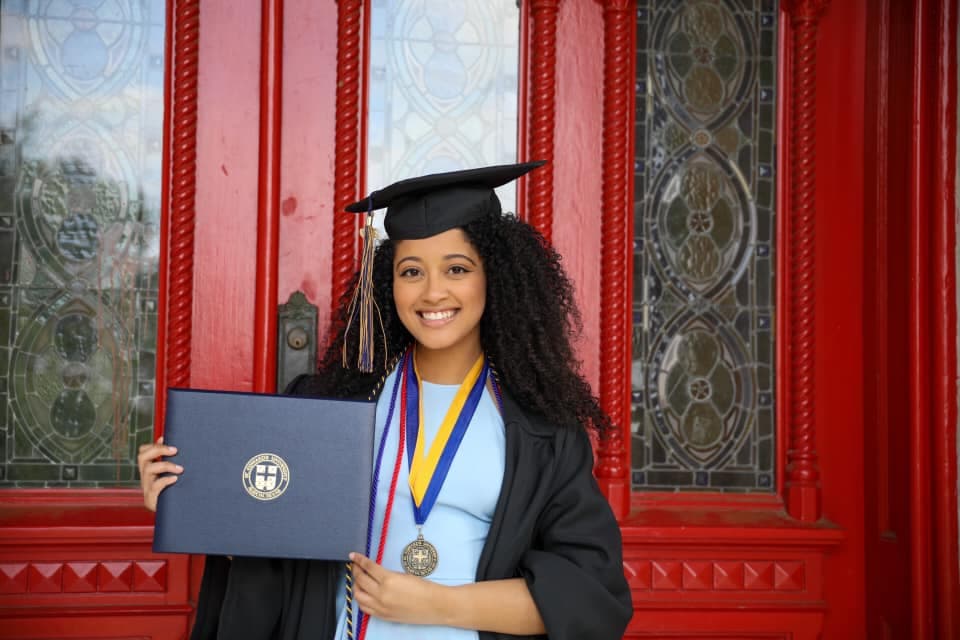 Smiling graduate in cap and gown holds her diploma before ornate red stained-glass doors.